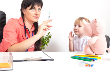 The doctor holds a medical syringe in his hands and a little girl cries nearby in fright. The...