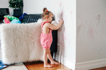 Funny cute baby girl drawing with marker on wall at home. Toddler girl child with milk bottle playing at home. Authentic candid childhood lifestyle moment. Young artist painting on wall at living room - 406739608