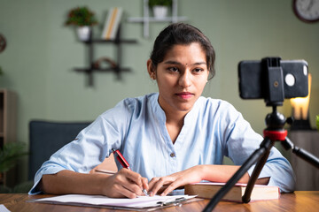 Young college girl student writing notes by looking into mobile phone during online Class - concept...