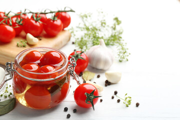 Glass jar of pickled cherry tomatoes and ingredients on white wooden table. Space for text