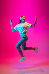 Fototapeta na wymiar Jumping high. Caucasian woman's portrait isolated on pink studio background in mixed neon light. Listening to music with phone. Concept of human emotions, facial expression, sales, ad, fashion