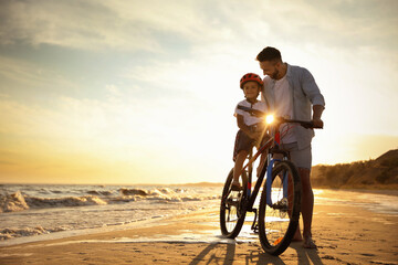 Happy father teaching son to ride bicycle on sandy beach near sea at sunset