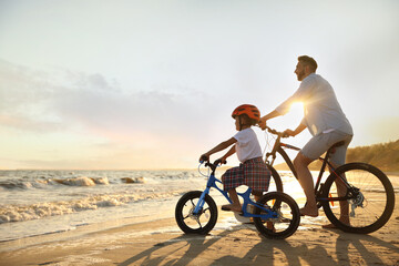 Fototapeta na wymiar Happy father with son riding bicycles on sandy beach near sea at sunset