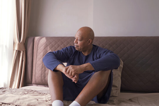 Depressed Lonely Old Elderly Black Man. African American People Sitting On Bed With Windows In Bedroom At Home In Early Morning. Quarantine Lifestyle. Unhappy Life.