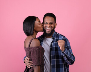Excited black guy making YES gesture, young girlfriend kissing his cheek on pink studio background. Valentine's Day