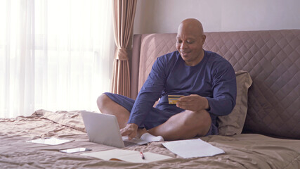 Happy old elderly black man. African American people shopping online, using a credit card to pay money with a computer notebook laptop, business documents on bed in bedroom at home. Lifestyle.