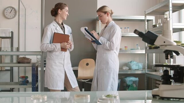 Lockdown of two female scientists wearing medical overall standing together in lab, communicating and making a report using modern devices