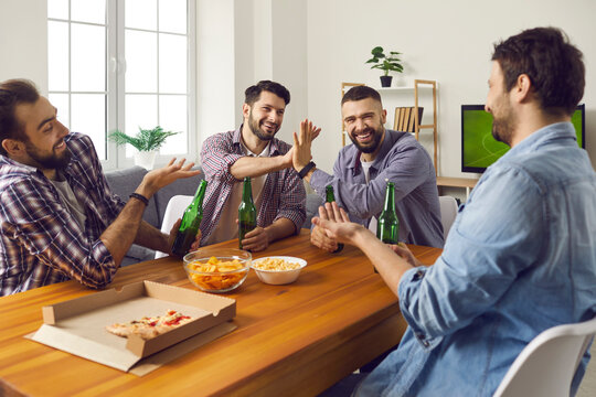 Male friends enjoying good time at fun get-together on weekend. Group of happy young men sitting at table at home, drinking beer, eating pizza, watching TV, playing games and telling funny stories