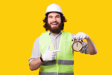 Photo of amazed bearded architect man wearing helmet and pointing at alarm clock.
