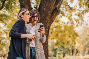Two happy smiling women friends checking smart phone content in a park