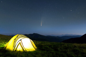 Tourist hikers tent in mountains at night with stars and Neowise comet with light tail in dark...