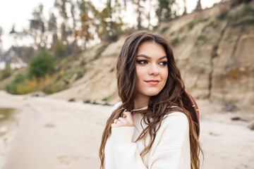 young beautiful girl in a white sweater, blue jeans and brown hat are walking on the coast near the lake
