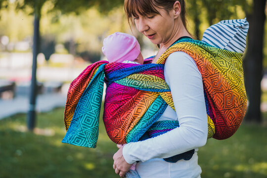 Beautiful Happy Young Mother With Baby Carrier In Park. Mom Walking With Infant In Baby Carrier. Happy Mother Carrying Her Child By Ergonomic Baby Carrier.