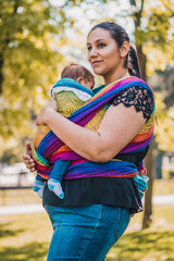 Beautiful happy young mother with baby carrier in park. Mom walking with infant in baby carrier. Happy mother carrying her child by ergonomic baby carrier.