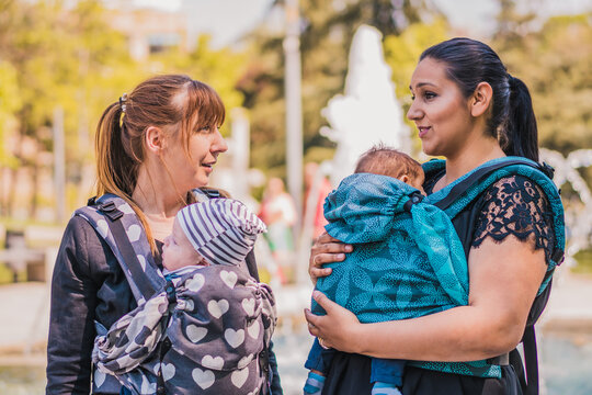 Two beautiful happy young mothers with baby carrier in park. Moms walking with infant in baby carrier. Happy mothers carrying her child by ergonomic baby carrier.