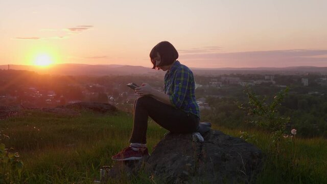 Side view of young white-skinned woman works using tablet computer, short-haired brunette sits on mountain rock at sunset. The concept of Internet availability regardless of location.