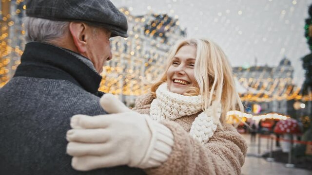 Happy senior blonde woman running to man, hugging him, enjoying Christmas meeting on city square