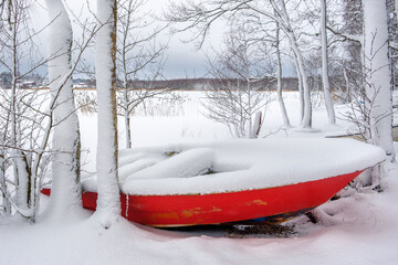 A red boat covered with snow on the snowy shore of the lake.