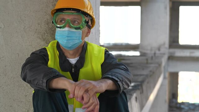 Worker in goggles, yellow hard hat and blue protective face mask sits at construction site and looks around. 4K 60fps tilt video