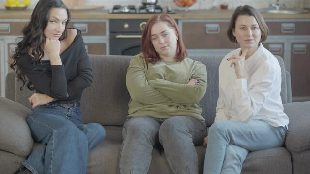Confident woman crossing hands and looking at camera as friends posing on both sides. Millennial Caucasian women posing indoors sitting on couch.