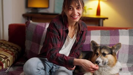 camera zooming on pretty smiling cute young woman sitting on couch with dog at evening. joyful girl watching film online at home and feeding her Welsh Corgi dog by own food