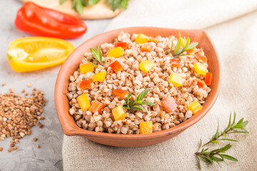 Buckwheat porridge with vegetables in clay bowl on a gray concrete background and linen textile. Side view, close up, selective focus.