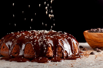 Side view of a fresh baked chocolate cake on white stone countertop with dark background; Melted chocolate pudding is dripping on the sides with coconut sprinkles falling from above for decoration.
