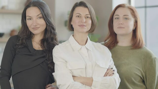 Three confident gorgeous young women posing indoors looking at camera. Portrait of beautiful Caucasian millennial ladies with serious facial expression.