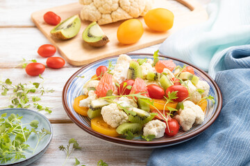 Vegetarian salad of cauliflower cabbage, kiwi, tomatoes, microgreen sprouts on gray wooden background. Side view, close up.