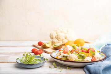 Vegetarian salad of cauliflower cabbage, kiwi, tomatoes, microgreen sprouts on gray wooden background. Side view, selective focus, copy space.