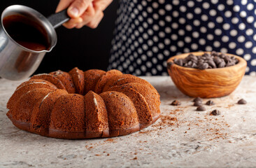A chef is about to pour melted chocolate on top of a round cake fresh out of oven. She is holding a metal pitcher which contains the melted icing. There are cacao sprinkles and a bowl of chocolate