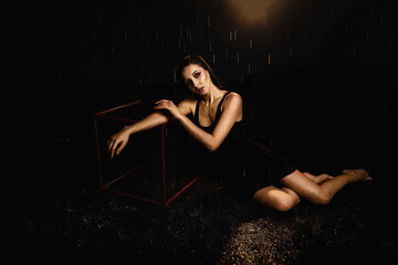 A slender brunette in a black dress on a black background in the studio under drops of rain, illuminated by spotlights