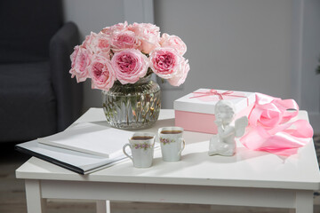 a bouquet of pink roses in a glass vase, two porcelain cups with tea, a white box with a heart with a gift, a satin ribbon, a figurine of an angel - gifts for Valentine's Day on a coffee table.