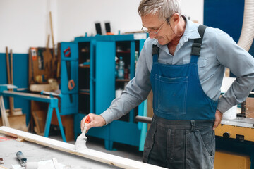 An elderly cabinetmaker in overalls and glasses paints a wooden board with a brush on a workbench in a carpentry shop. Non-staged photo while working with copy space