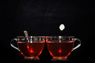 A piece of rock sugar is splashing into one of two glass cups of tea in front of a black background