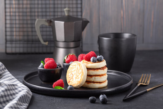 Cottage cheese pancakes with fresh with fresh raspberries and blueberries on dark ceramic plate. Cup of coffee and coffee maker - Image