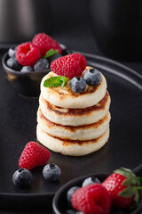 Close up of cheese pancakes with fresh berries on dark ceramic plate. Syrniki. Vertical photo - Image