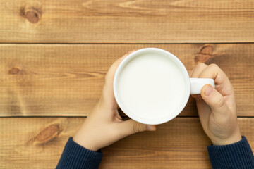 Child hands holding cup of milk against blurred wooden background, top view. Farm dairy products concept.
