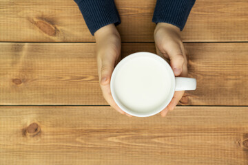 Child hands holding cup of milk on wooden table, top view. Farm dairy products concept.