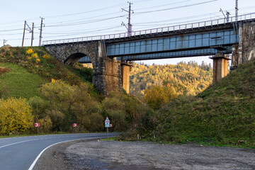 An old stone arched railway bridge over a road in the mountainous part of the Ukrainian Carpathians. Autumn mountain landscape - yellowed and reddened autumn trees combined with green needles.