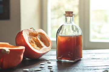 A bottle of organic pumpkin seed oil and pieces of fresh pumpkin on a wooden table against a window