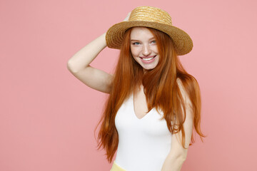 Young smiling atractive cute redhead woman 20s ginger long hair wearing straw hat summer clothes looking camera playing put hand on head isolated on pastel pink color background studio portrait.