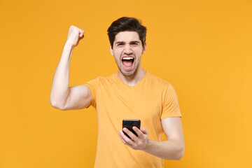 Young shocked surprised unshaved man 20s in casual basic blank print design t-shirt holding modern mobile cell phone doing winner gesture saying yes isolated on yellow background studio portrait.