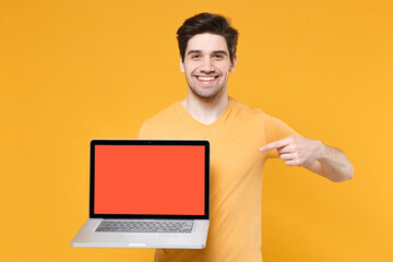 Young smiling unshaved man in basic print design t-shirt using pointing index finger on laptop pc computer with blank screen workspace copy space mock up isolated on yellow background studio portrait.