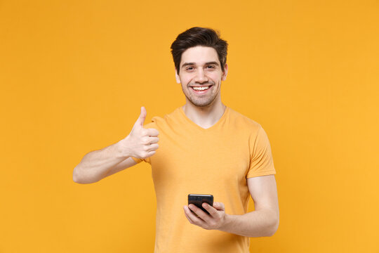 Young smiling unshaved caucasian satisfied man 20s in casual basic blank print design t-shirt holding modern mobile cell phone showing thumb up isolated on yellow color background studio portrait