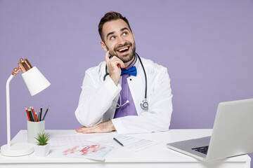Dreamful pensive male doctor man in white medical gown suit sit at desk work on computer documents...