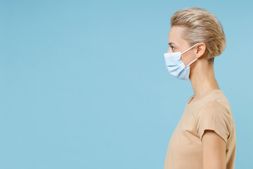 Side view of young blonde woman 20s short haircut in beige t-shirt sterile face mask to safe from coronavirus virus covid-19 pandemic quarantine look aside isolated on blue background studio portrait.