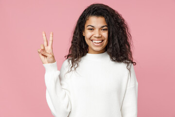 Young happy positive cheerful cute african american woman 20s curly hair in white knitted sweater showing victory v-sign gesture isolated on pastel pink background studio portrait Lifestyle concept.