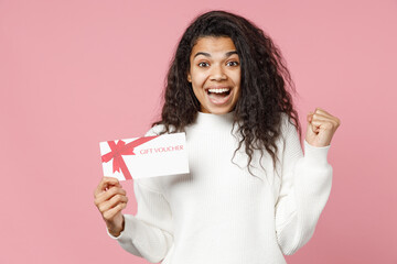 Young happy african american woman 20s curly hair in white casual knitted sweater holding gift voucher flyer mock up do winner gesture clenhing fist isolated on pastel pink background studio portrait