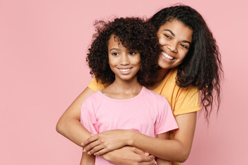 Cheerful pretty african american young woman and little kid girl sisters wearing casual t-shirts hugging looking aside isolated on pastel pink color background studio portrait. Family day concept.